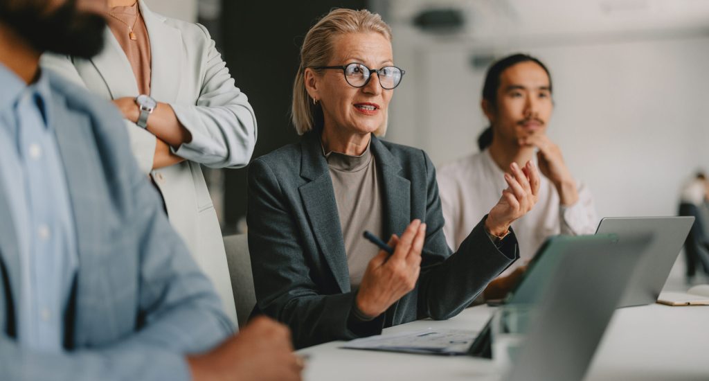 A diverse group of professional individuals engaged in a business meeting within a bright beige office, illustrating teamwork, collaboration, and leadership.